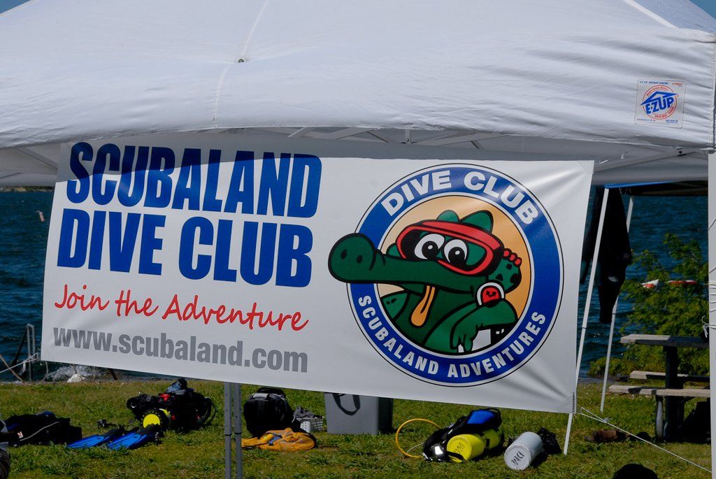Scubaland Dive Club banner with cartoon alligator. Equipment sits in front of a white tent on a grassy area near water.