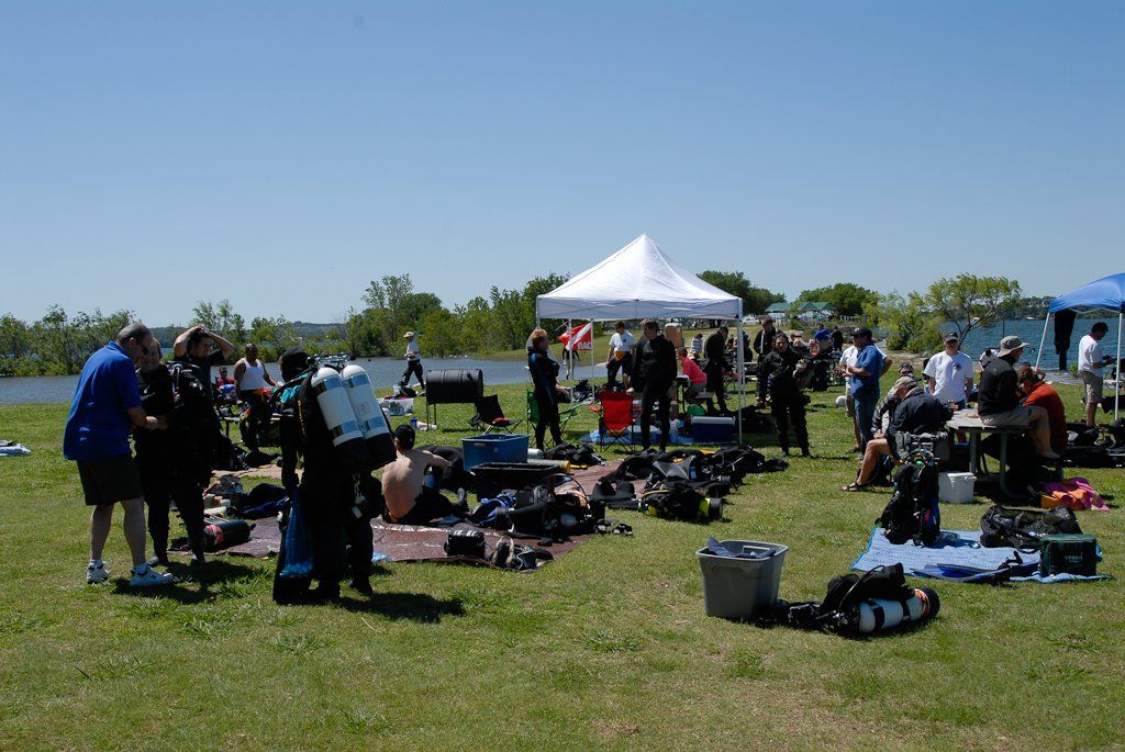 Divers preparing equipment on grassy shore, white tent in background, water and trees visible.