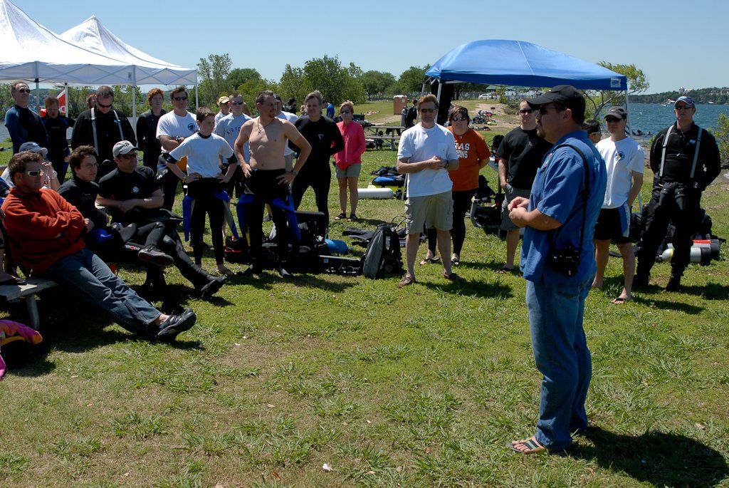 A man speaks to a group of people outdoors. Some wear wetsuits, and tents are in the background.