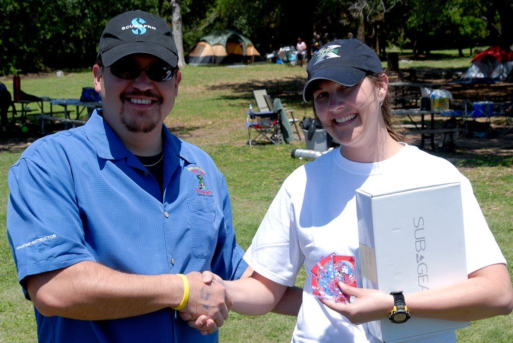 Man in blue shirt shakes hands with woman in white, holding a box. Outdoors, grass, tents in background.