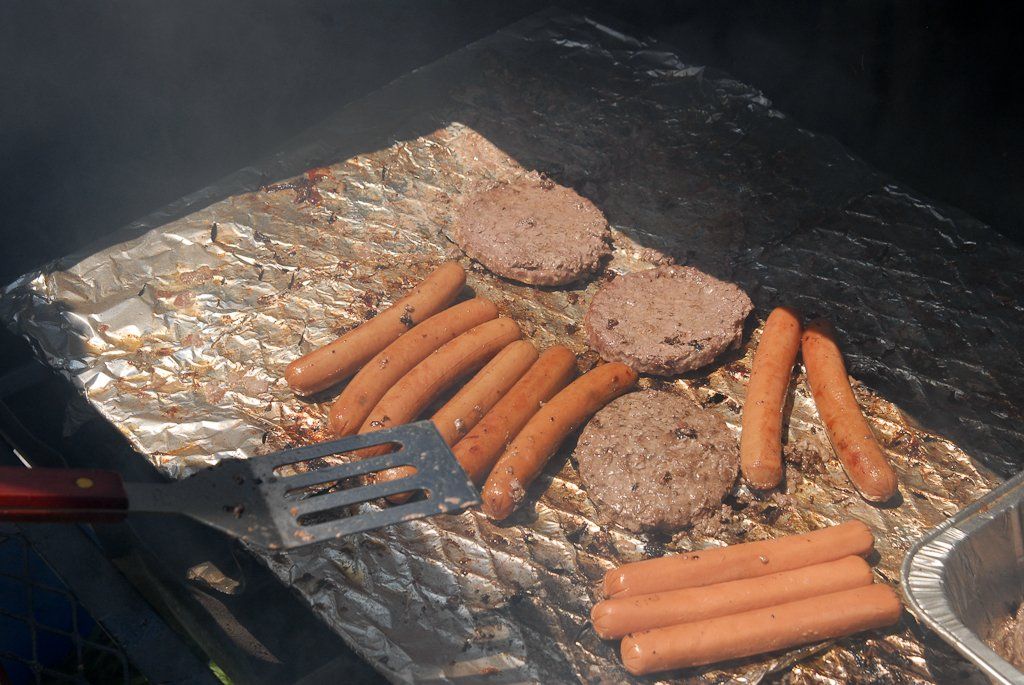 Hotdogs and hamburgers cooking on foil-lined grill with a spatula.