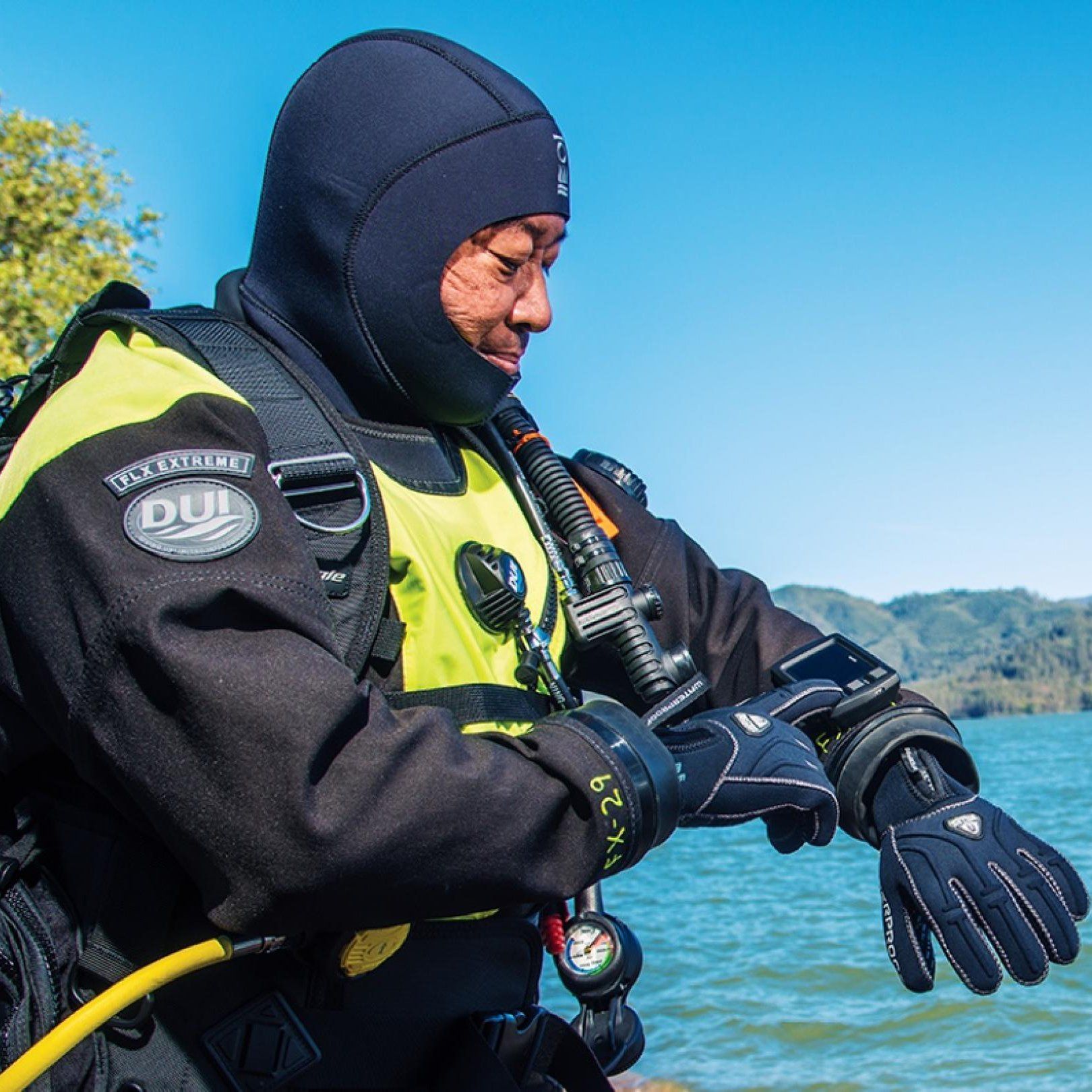 Diver in black and yellow drysuit checks their watch near a body of water.