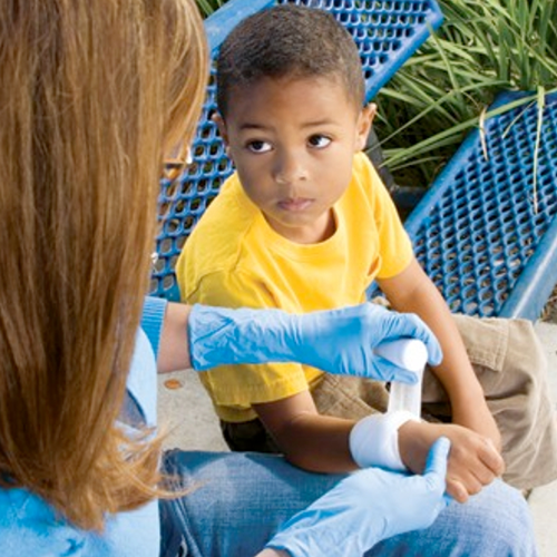 Person bandaging a child's wrist outdoors. Child in yellow shirt looks on.