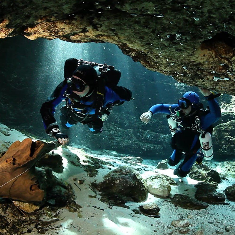 Two scuba divers explore a sunlit underwater cave; one touches the wall, while the other floats by.