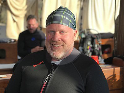 Man wearing a wetsuit and plaid head covering, smiling, in front of scuba gear.
