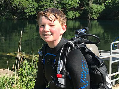 Boy in wetsuit with scuba gear smiling by a lake.