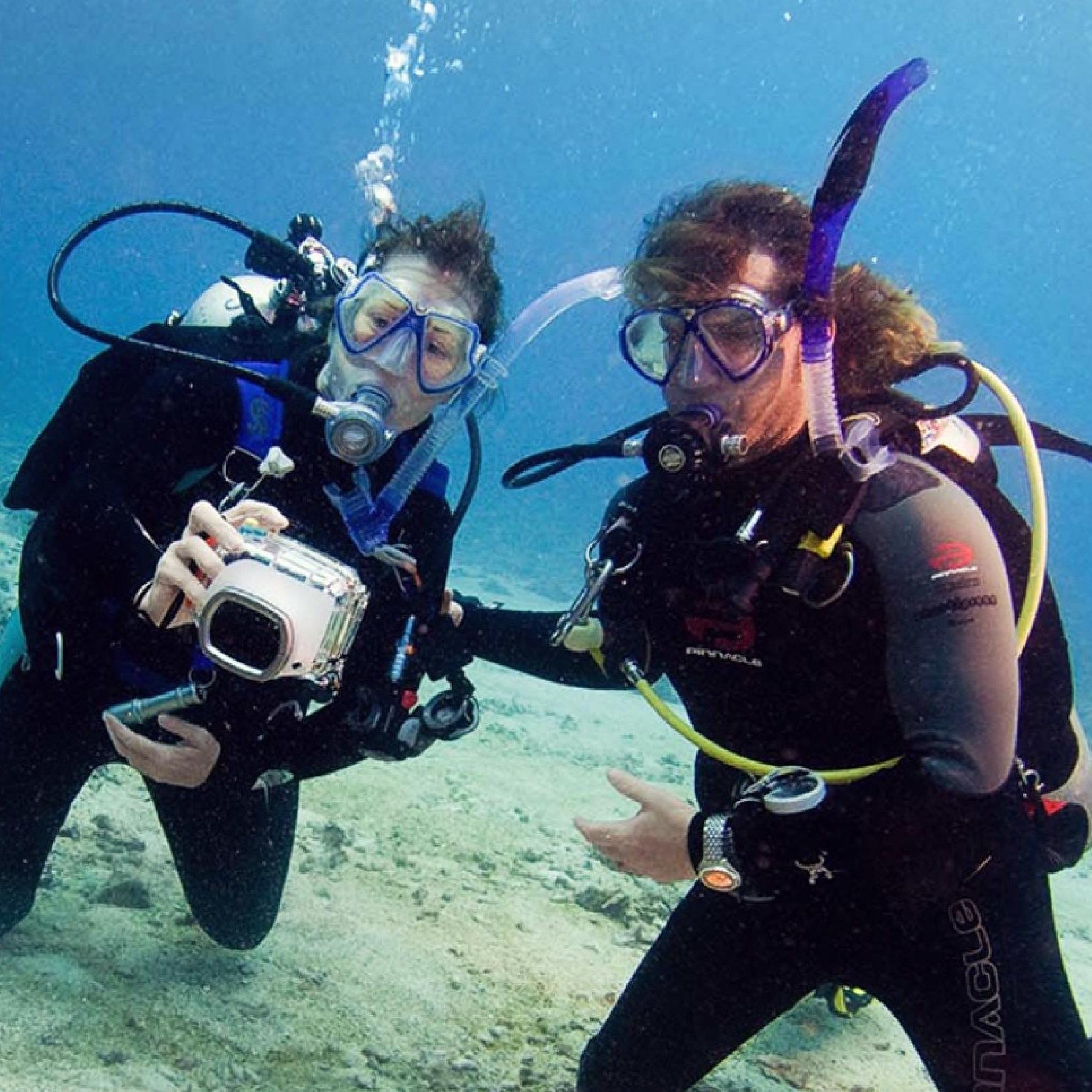 Two divers underwater, one holding camera. Both wear wetsuits, masks, and scuba gear in clear blue water.