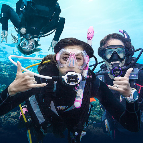 Three scuba divers underwater, smiling, giving hand gestures. Pink and black dive gear.
