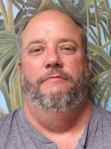 Man with a gray beard and shirt; neutral expression; background with green and blue plants.