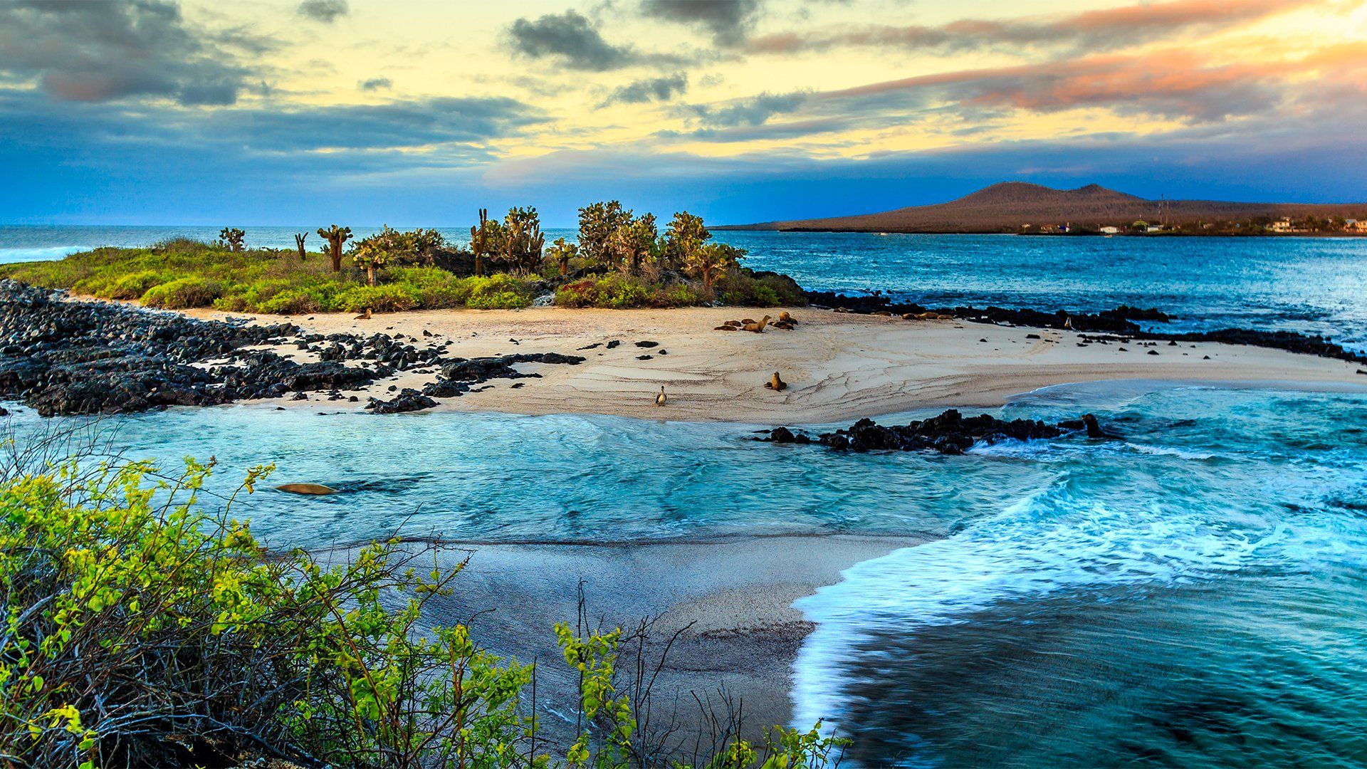 Island beach with turquoise water, rocky shore, and distant volcanic island under a colorful sky.