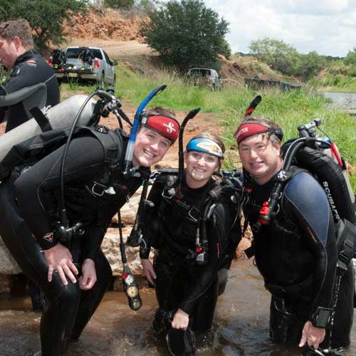 Divers in wetsuits standing in water, smiles, ready to dive.