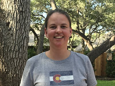 Woman in gray Colorado flag t-shirt smiles in front of a tree outdoors.