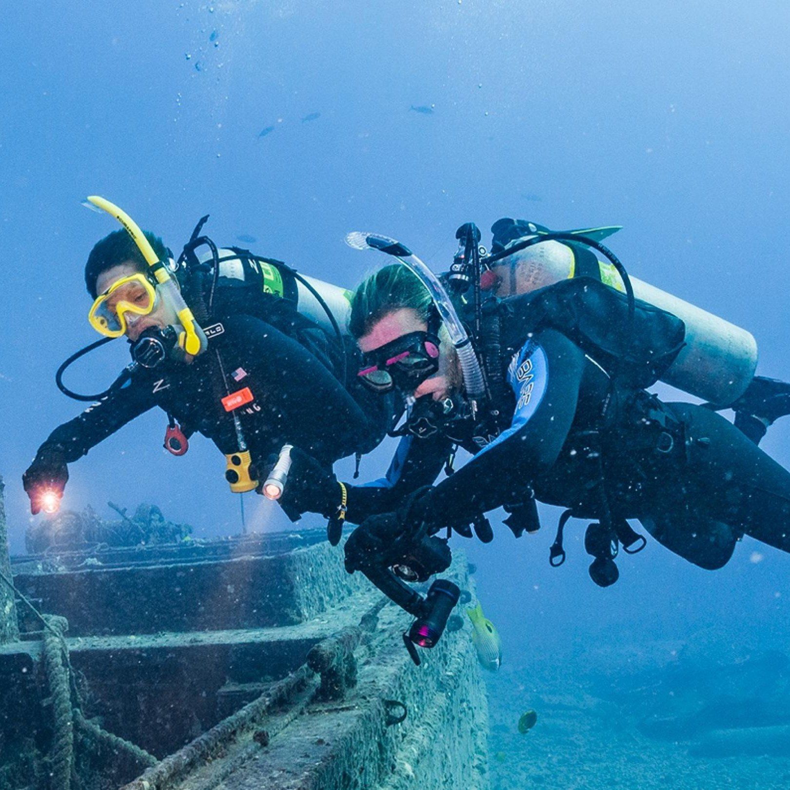 Two scuba divers explore a sunken shipwreck, illuminated by underwater lights.