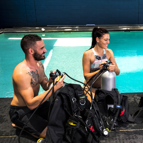 Man and woman in scuba gear preparing to dive in an indoor pool.