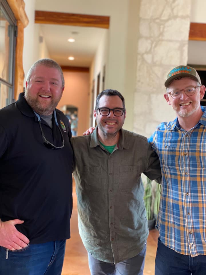 Three men smiling, arm-in-arm in a hallway. One in a black polo, one in a green shirt, and one in a plaid shirt with a hat.