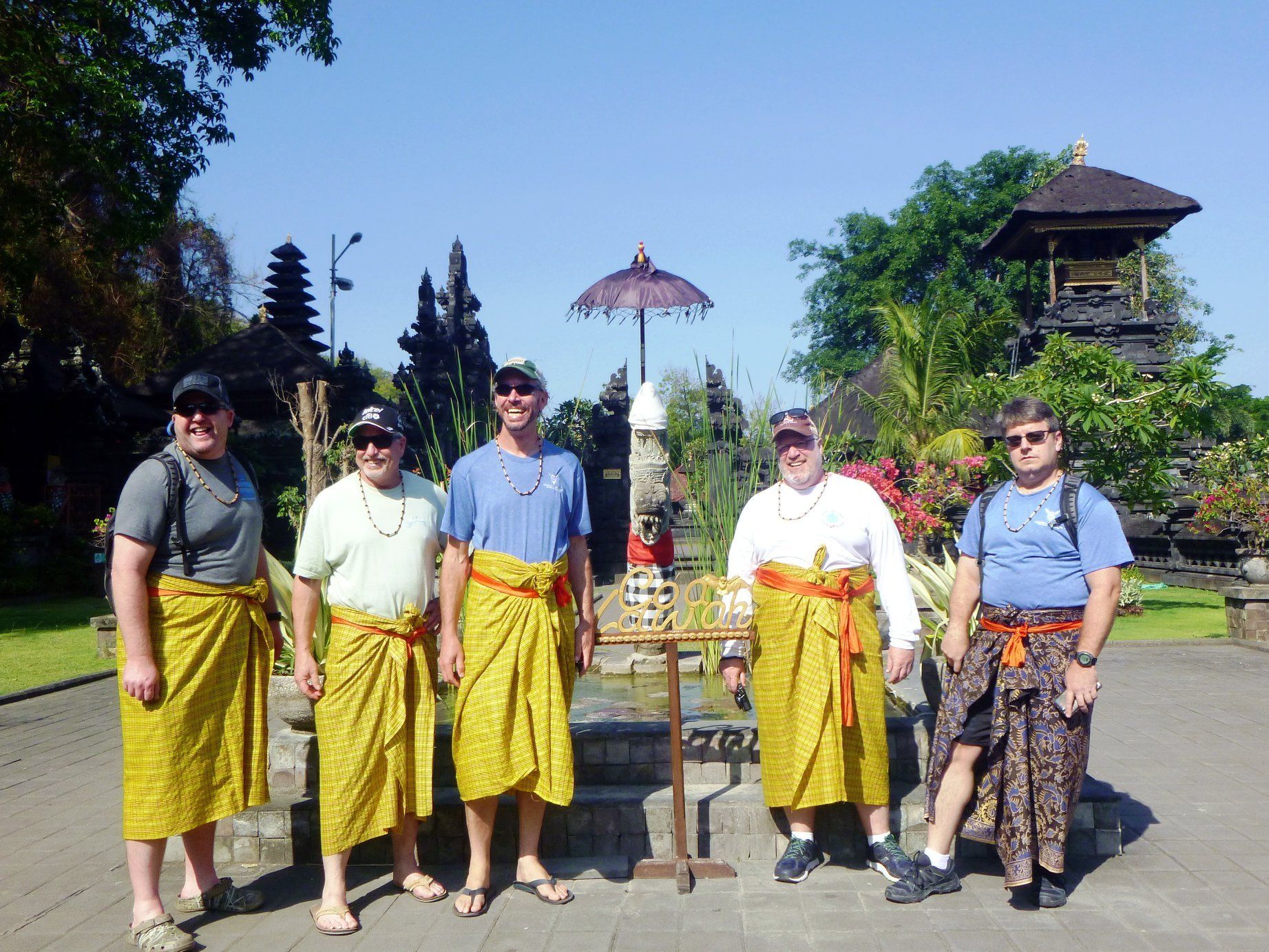 Five people wearing sarongs pose at a temple. Bright yellow sarongs, blue sky, and green trees are visible.