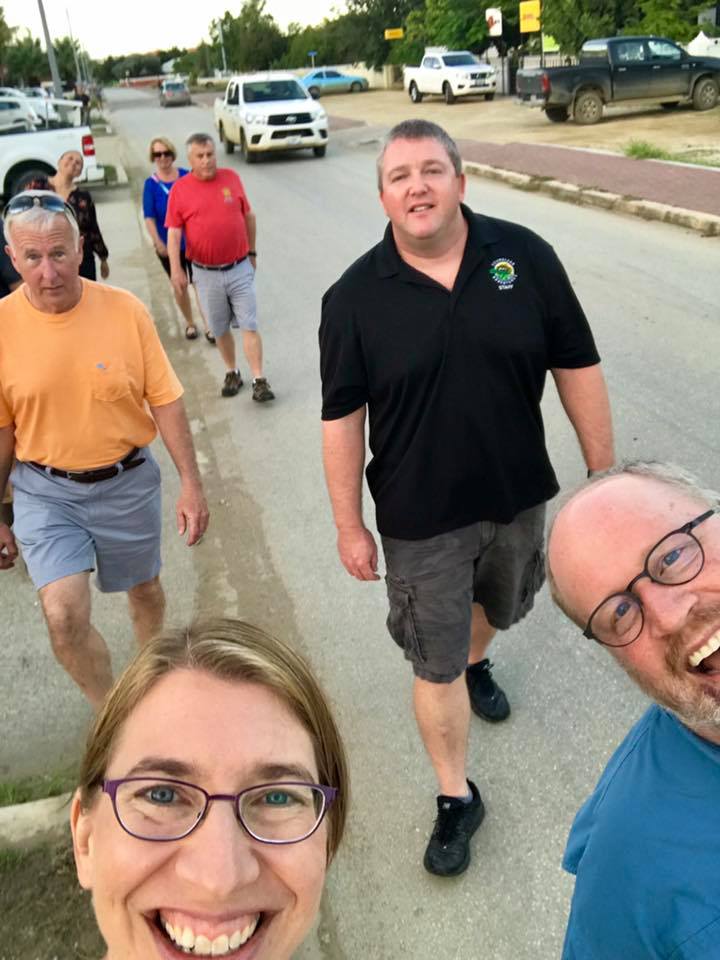 Group of people walking on a road; some smiling. Bright day with vehicles and buildings in the background.