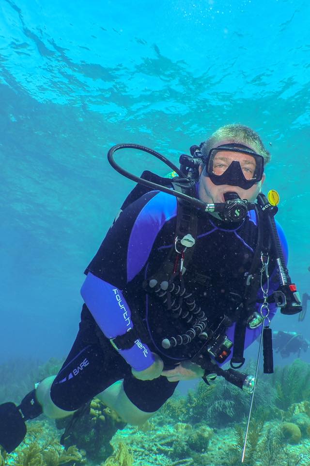 Scuba diver in blue and black wetsuit under water, near coral, looking at camera.
