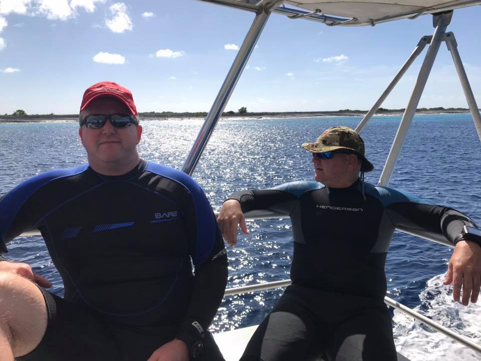 Two men in wetsuits on a boat, bright sun reflecting on the blue water.
