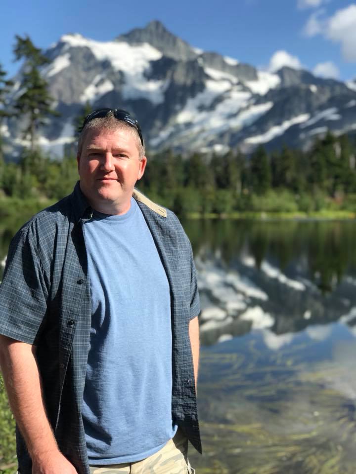 Man in blue shirt and jacket stands near a lake, with snow-capped mountains reflected in the water.
