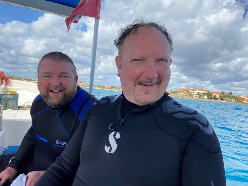 Two people on a boat in water, wearing wetsuits. One with a smile, the other looking at the camera.