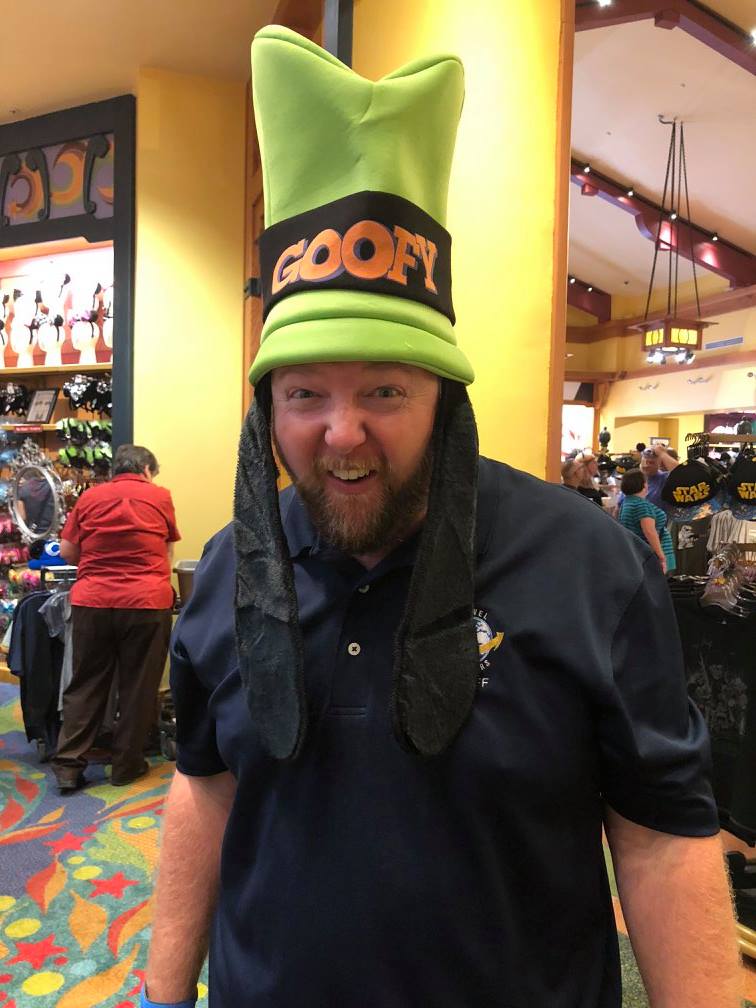 Man wearing a Goofy hat inside a store, smiling. Green and black hat, blue shirt.