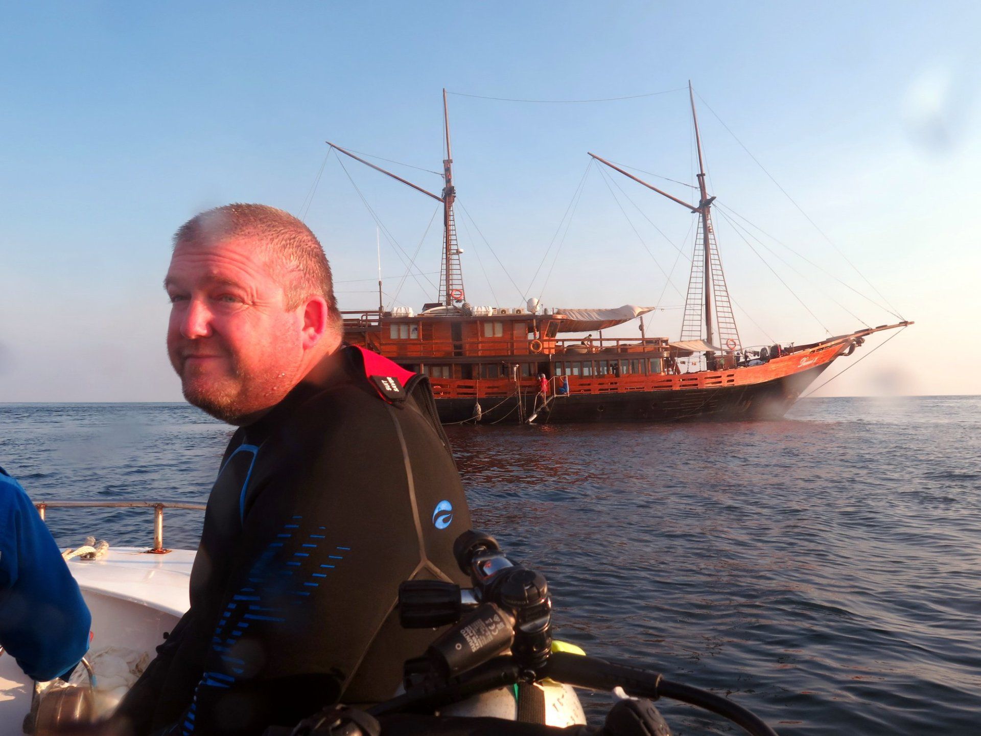 Man in dive gear on boat with large wooden sailboat in background. Blue water.