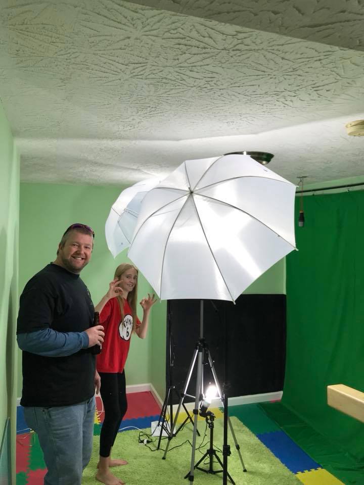 Man and girl smiling, posing in front of a green screen. They are in a room with a light and umbrella setup.