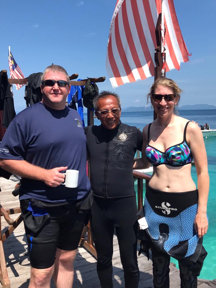Three people pose on a wooden dock with blue water and flags. They are smiling, two in diving gear.