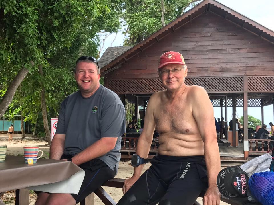 Two men sitting on a bench near a beach hut. One man shirtless, in swim gear. The other wears a t-shirt.