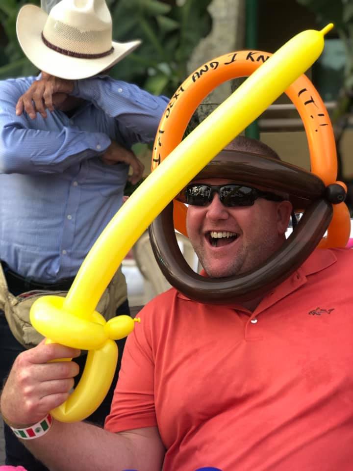 Man wearing balloon hat laughs, holding a yellow balloon sword; cowboy in background.