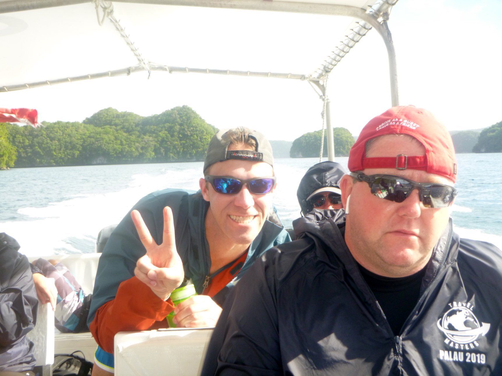 People on a boat near a tropical island, one giving a peace sign, all wearing sunglasses.