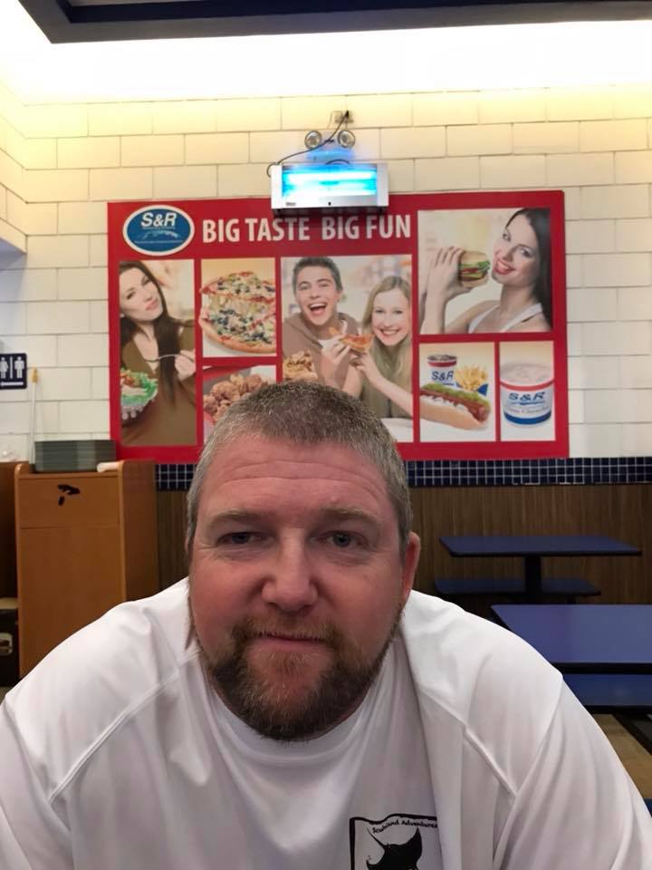 Man in white shirt smiling in front of a colorful restaurant sign that says
