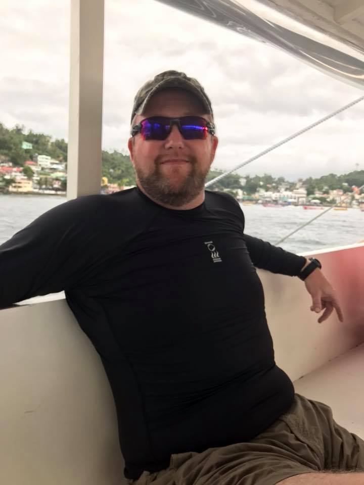 Man wearing sunglasses and a black long-sleeved shirt, sitting on a boat with a coastal town in the background.