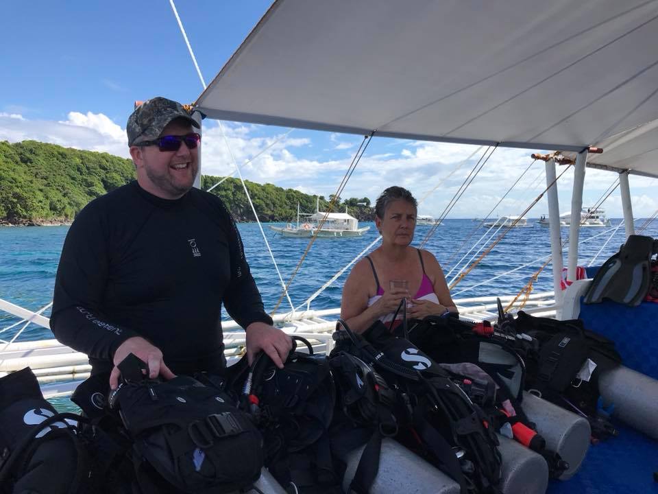 Man and woman on a boat, surrounded by scuba gear, smiling at the camera.