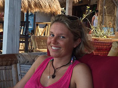 Woman with blonde hair wearing a pink tank top smiles outdoors in a tropical setting.