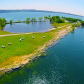 Lakeside park with green grass, picnic tables, trees, and calm, blue water under a clear sky.