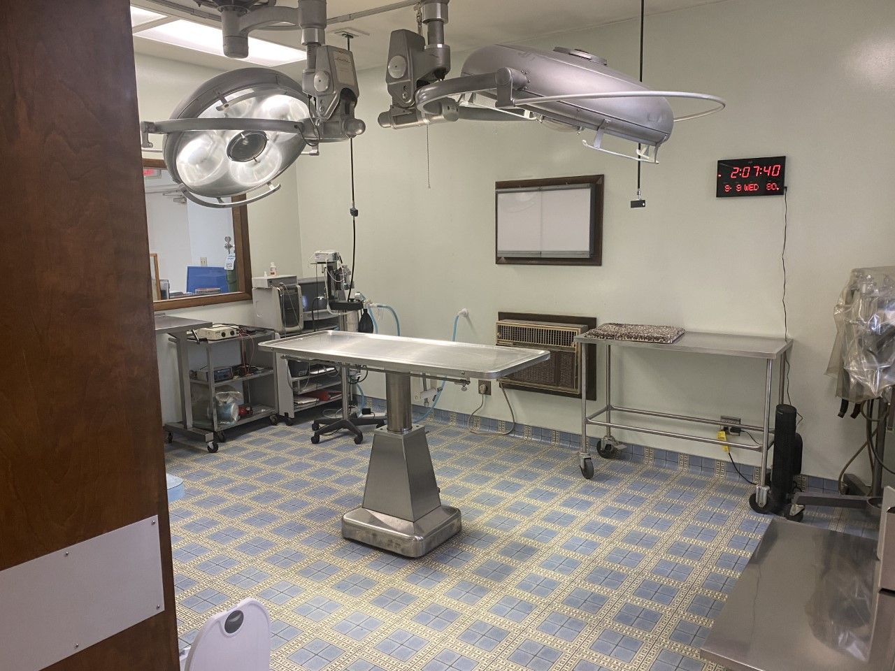 Operating room with surgical lights, stainless steel table, and medical equipment. Blue tiled floor.