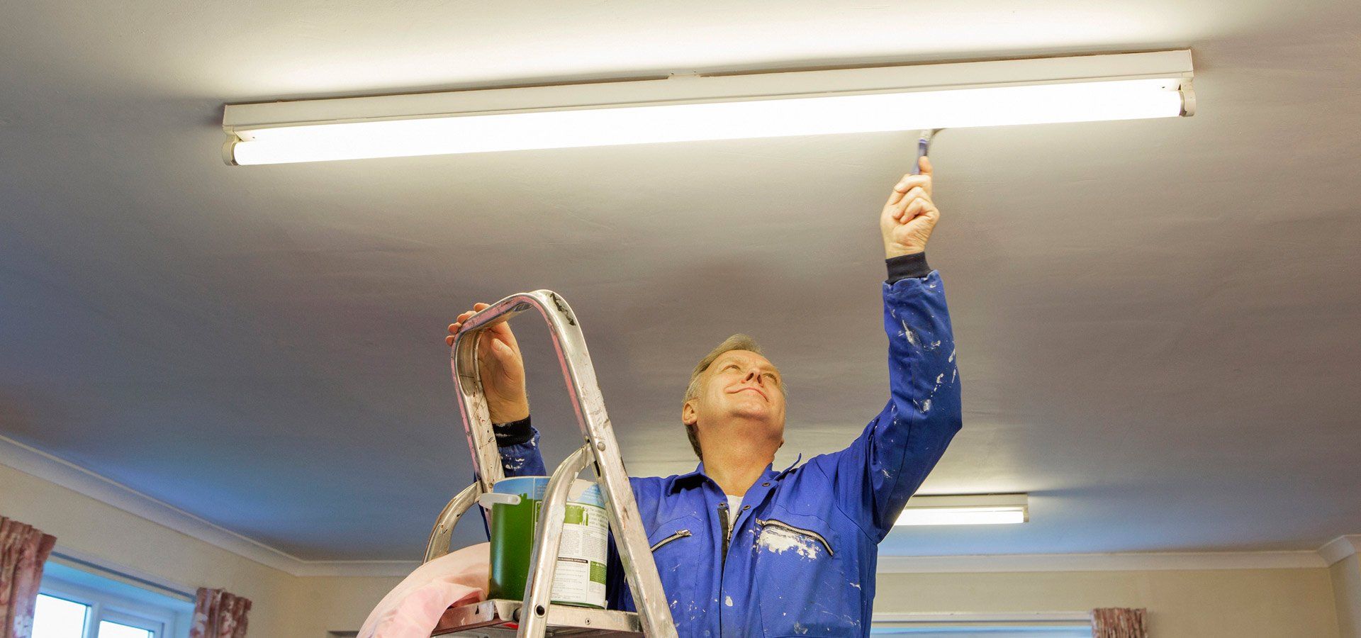 A person on a ladder, painting a ceiling. They are wearing a blue jumpsuit, in a room with a light fixture.