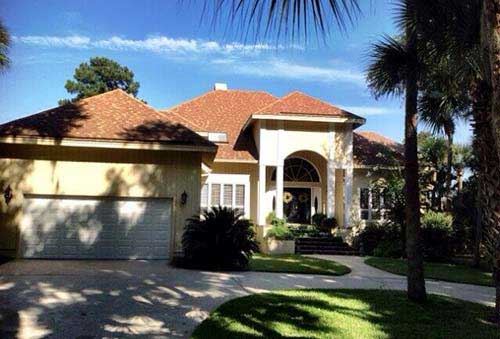 Residential Roofing — House with Shingles Roof in Savannah, GA Beige stucco house with brown roof and palm trees, driveway and grass in front.