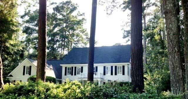 Master Elite Contractor — GAF Roofing in Savannah, GA White house with dark shutters and roof, nestled among trees.
