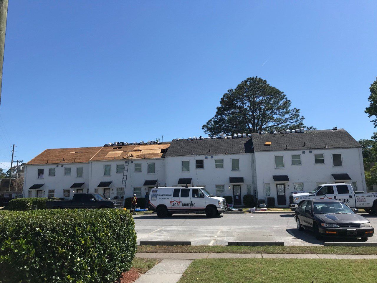 Workers repairing a white apartment building's roof under a clear, sunny sky; several vans parked in the parking lot.
