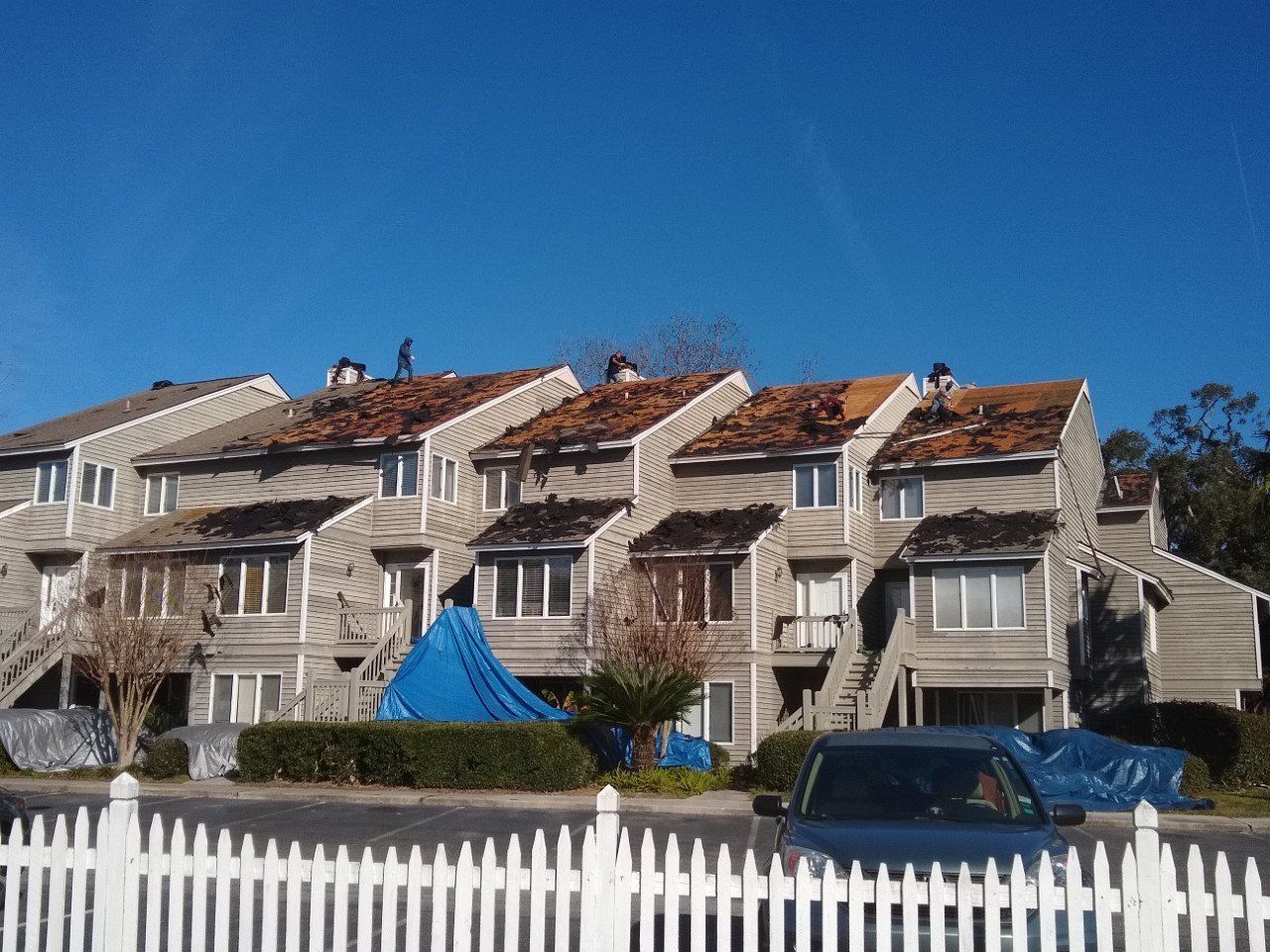 Townhouses with roofing work in progress, blue tarps covering areas, bright blue sky.
