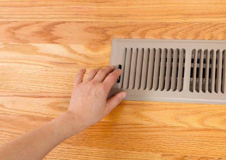 A hand adjusting a beige metal floor air vent register installed in a light-colored hardwood floor.