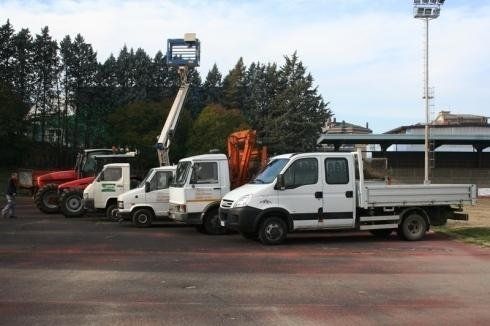 Una fila di veicoli di manutenzione parcheggiati, tra cui un trattore rosso e diversi camion bianchi, uno dei quali dotato di piattaforma aerea sopraelevata.