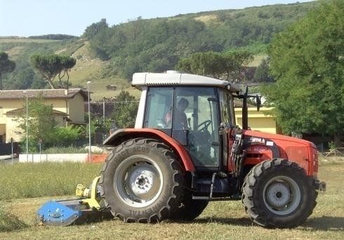 Un trattore rosso con un tosaerba blu al lavoro in un campo erboso di fronte ad alcuni edifici e a una collina.