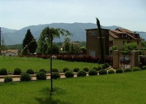 Un prato verde con arbusti curati conduce a una casa con il tetto di tegole, incorniciata da alberi e montagne in lontananza sotto un cielo azzurro.