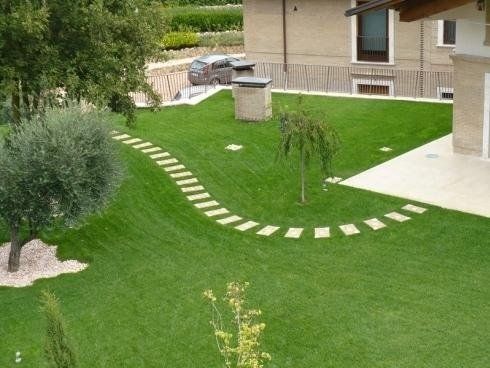 Un sentiero in pietra si snoda attraverso un prato verde ben curato di fronte a una casa, dove si trovano un ulivo e un piccolo albero ornamentale.