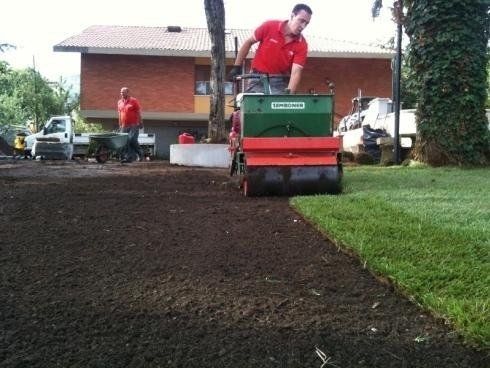 Una persona con una maglietta rossa aziona un rullo verde e rosso per stendere un nuovo prato davanti a una casa in mattoni.