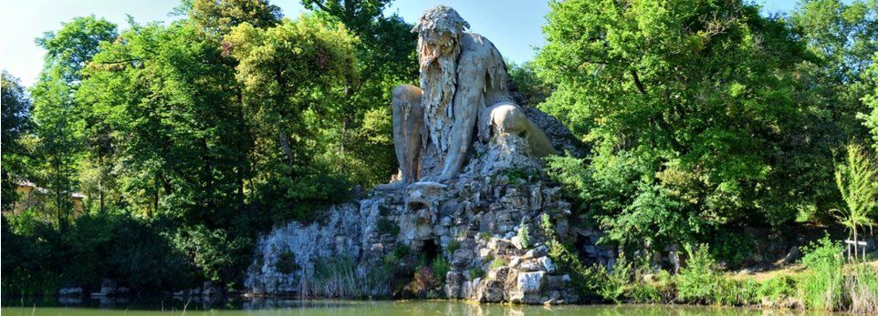 Una colossale statua di pietra raffigurante un uomo barbuto è accovacciata su una grotta rocciosa accanto a uno stagno, circondata da rigogliosi alberi verdi.